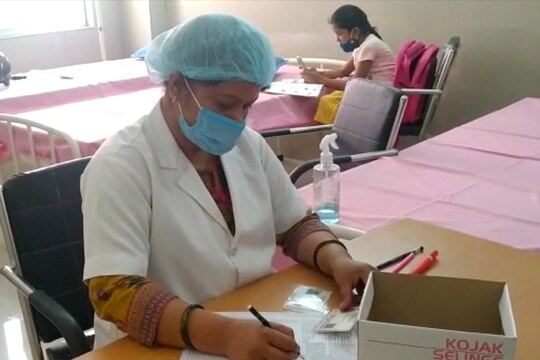 आप देख सकतें हैं वैक्सीनेशन कमरे में एक तरफ नर्स किरण हैं और उनके पीछे पढ़ाई करती उनकी बच्ची.