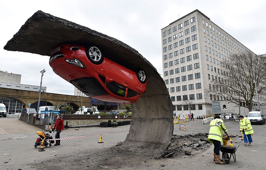 Pedestrians and workers pass an upside-down car art installation in a car park on the South Bank in London. British artist Alex Chinneck's illusory piece, entitled