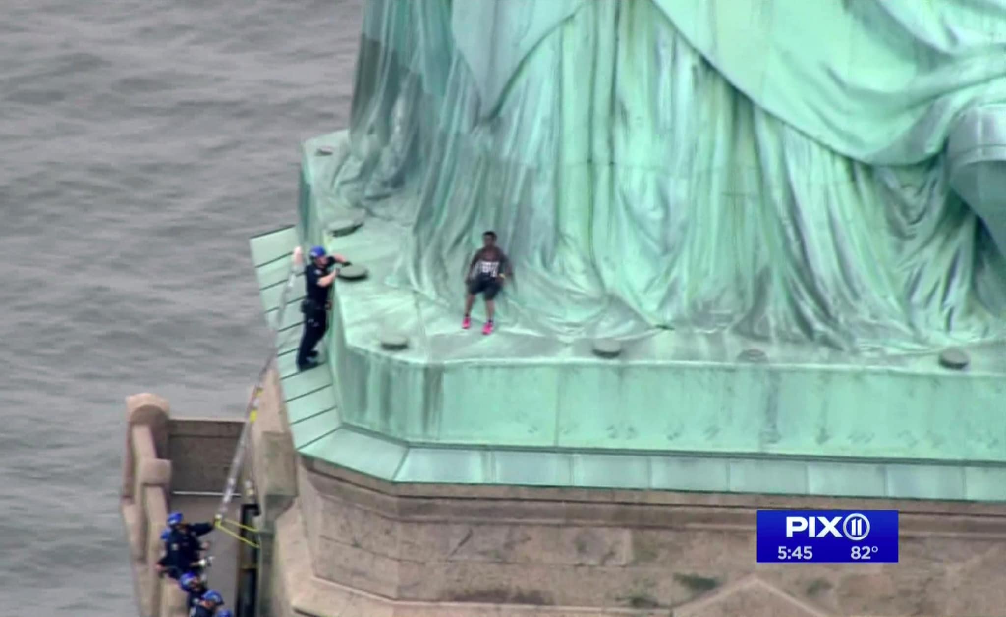 In this image made from video by PIX11, a person, center, leans against the robes of the Statue of Liberty on Liberty Island, as one of the police officers climbed up on a ladder to stand on a ledge nearby talking the climber into descending in New York, Wednesday, July 4, 2018. (PIX11 via AP)