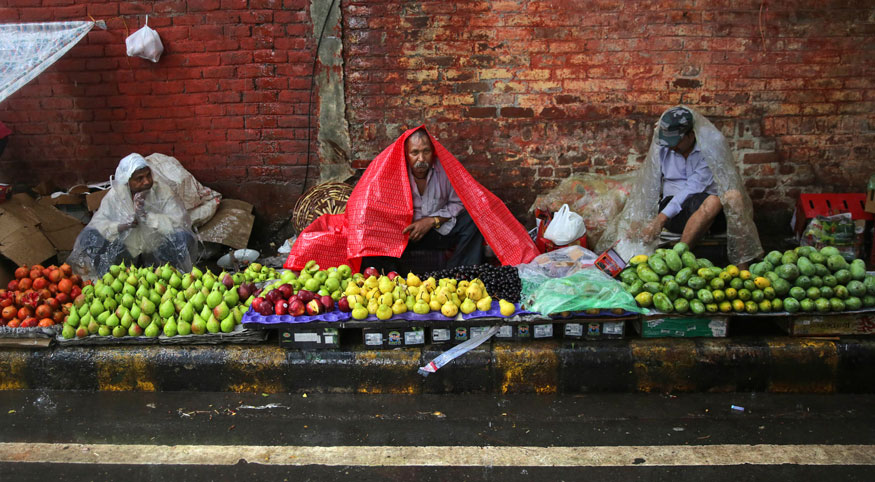 Roadside fruit vendors cover themselves with plastic sheets as they wait for customers amidst rain in New Delhi. (Image: AP)