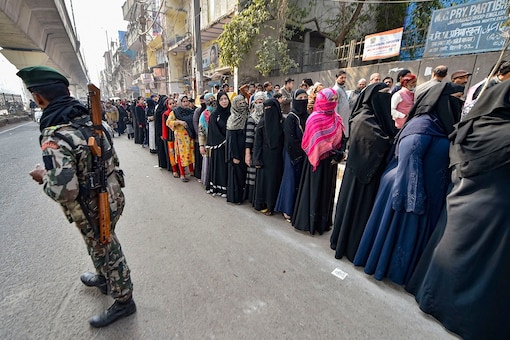 A security personnel stands guard as voters stand in a queue to cast vote during the Delhi Assembly elections at a polling station, in Jafrabad area of Delhi, Saturday, February 8, 2020. (Image: PTI)