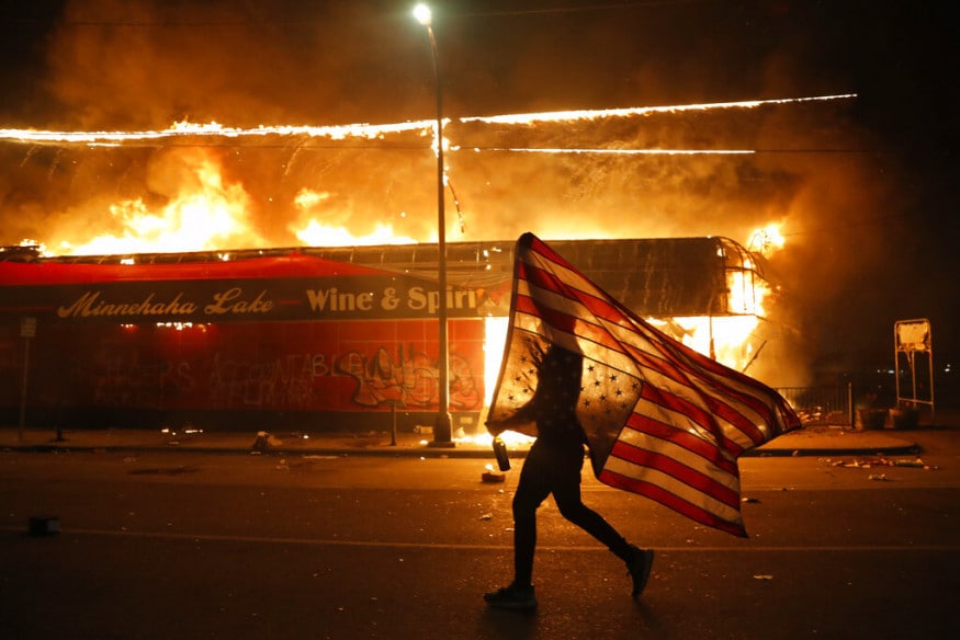  A protester carries a US flag upside down, a sign of distress, next to a burning building, in Minneapolis. (Image: AP)