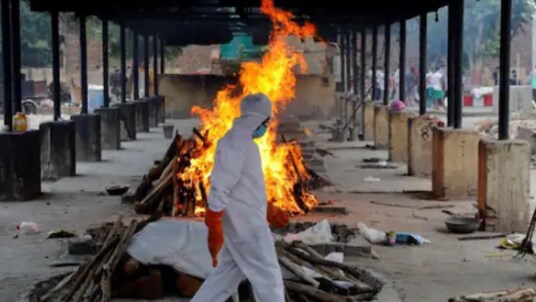 A priest wearing personal protective equipment (PPE) walks in front of the body of a person who died of the coronavirus disease (COVID-19), as he collects woods to make a funeral pyre at a crematorium in New Delhi, India, July 3, 2020. REUTERS/Anushree Fadnavis.

