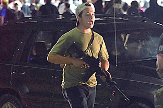 FILE - In this Tuesday, Aug. 25, 2020 file photo, Kyle Rittenhouse carries a weapon as he walks along Sheridan Road in Kenosha, Wis., during a night of unrest following the weekend police shooting of Jacob Blake. The way his lawyers tell it, the teenager wasn't a scared, gun enthusiast in over his head when he fatally shot two protesters. He was a courageous defender of liberty, a patriot exercising his right to bear arms amid chaos in the streets. But some legal experts say Rittenhouse's lawyers are taking big risks by turning a fairly straightforward self-defense case into a sweeping political argument that mirrors the law-and-order re-election campaign of President Donald Trump. (Adam Rogan/The Journal Times via AP)