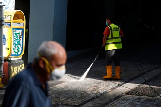 A municipal worker sprays disinfectant on a pavement as a man wearing face mask to prevent the spread of the new coronavirus walks at Ermou Street, Athens' main shopping area, Monday, Sept. 21, 2020. Greece is tightening restrictions in the greater Athens region, stepping up testing and creating quarantine hotels due to an increase in COVID-19 infections after Athenians returned from their summer holidays. (AP Photo/Thanassis Stavrakis)