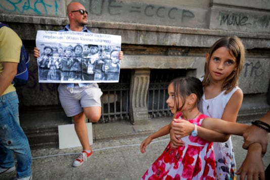 Children play next to a man holding a banner showing children in a concentration camp and a paraphrase from a statement by Romanian President Klaus Iohannis that reads 