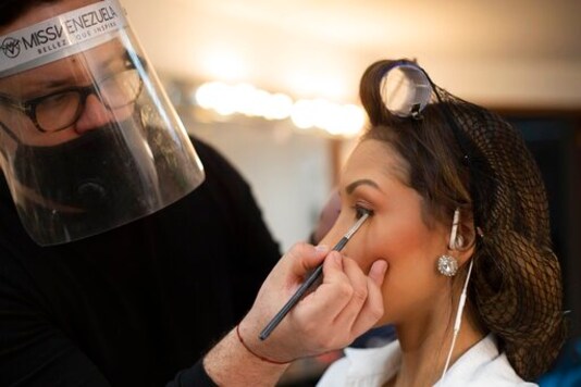 Stylist Juan Bautista, wearing a protective face mask and shield, applies eyeliner to Lara state beauty pageant contestant Jhosskaren Carrizo, in a Venevision television station dressing room, before a meeting with judges, in Caracas, Venezuela, Friday, Sept. 18, 2020. Quarantine rules and social distancing has forced the contestants to train at home and online with limited access to the venue itself where strict measures are in place. The pageant is due to take place on Sept. 24. (AP Photo/Ariana Cubillos)
