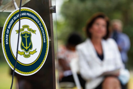 FILE - In this Monday, June 22, 2020 file photo, the seal of the State of Rhode Island decorates a podium as Gov. Gina Raimondo looks on at right during a news conference where she announced that she has signed an executive order to remove the phrase FILE - In this Monday, June 22, 2020 file photo, the seal of the State of Rhode Island decorates a podium as Gov. Gina Raimondo looks on at right during a news conference where she announced that she has signed an executive order to remove the phrase