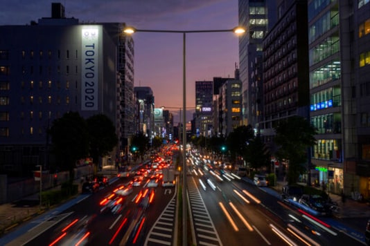 This long exposure photo shows streaks of lights from the cars passing by a Tokyo 2020 Olympics and Paralympics sign on the side of a building Monday, Oct. 12, 2020, in Tokyo, as the sky is colored by the sunset. (AP Photo/Kiichiro Sato) This long exposure photo shows streaks of lights from the cars passing by a Tokyo 2020 Olympics and Paralympics sign on the side of a building Monday, Oct. 12, 2020, in Tokyo, as the sky is colored by the sunset. (AP Photo/Kiichiro Sato)