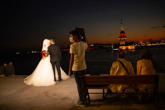 A wedding photographer's assistant, wearing a mask for protection against the spread of coronavirus, holds lighting equipment, as a couple poses for photographs backdropped by Istanbul's iconic Maiden's Tower (Kiz Kulesi) at the Bosphorus Strait separating the European and Asian sides of Istanbul, Friday, Sept. 11, 2020. Turkey is getting tough on people who flout self-isolation rules despite testing positive for the coronavirus. An Interior Ministry circular sent to the country's 81 provinces on Friday said people caught leaving their homes despite isolation orders will be quarantined and supervised at state-owned dormitories or hostels. (AP Photo/Yasin Akgul)