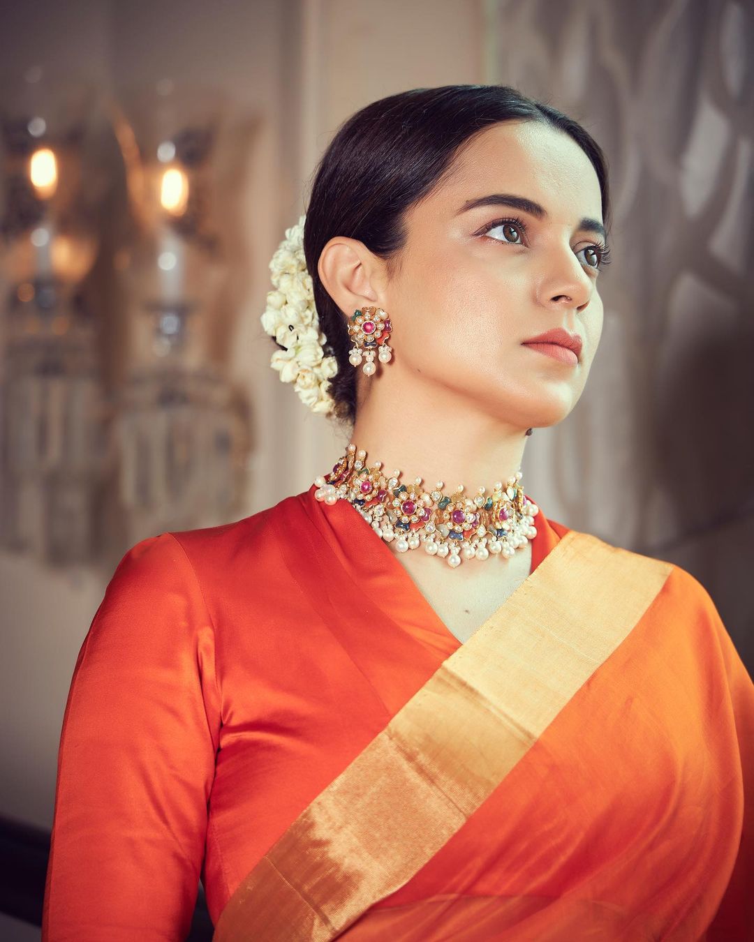Kangana Ranaut pairs her orange saree with a choker and fresh flowers adorn her hair. 
