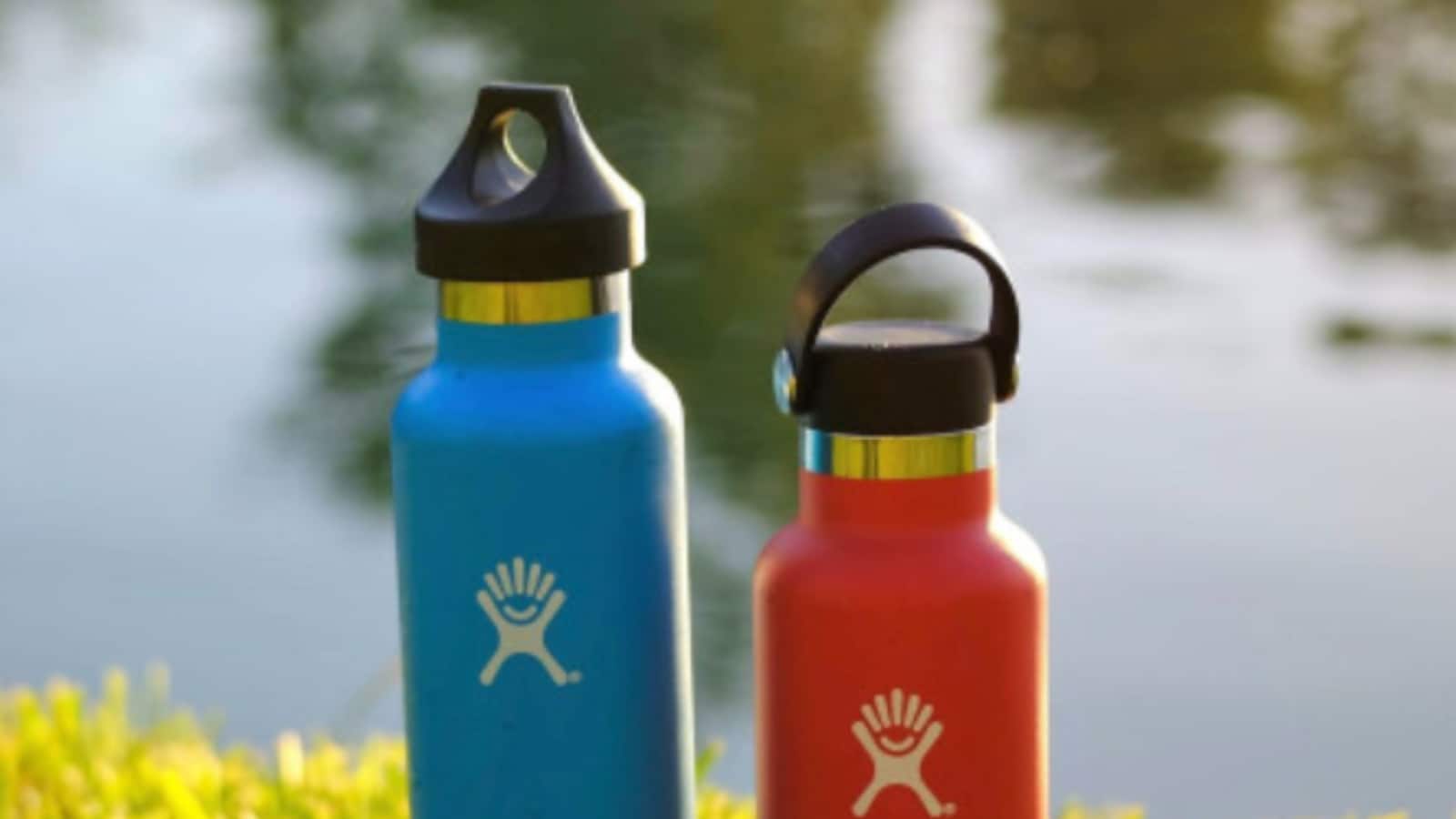  Two Hydro Flask water bottles in blue and red colors are sitting on the green grass near the lake.