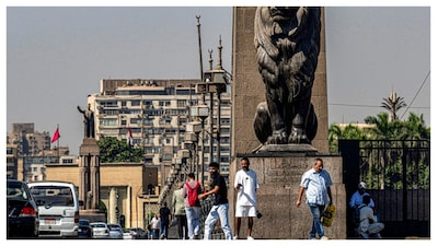 People walk past one of the four bronze lion statues at the end of the landmark Qasr el-Nile bridge connecting Cairo's central Tahrir Square to the island of Zamalek (background) on October 6, 2025 (AFP)